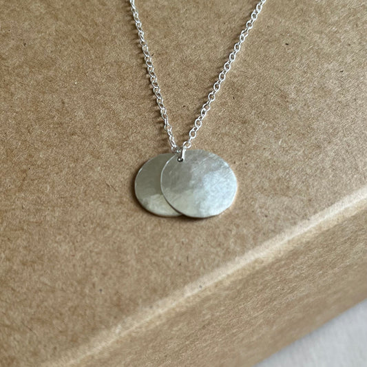 Close up image of silver double disk necklace on silver chain shown on a Kraft gift box.